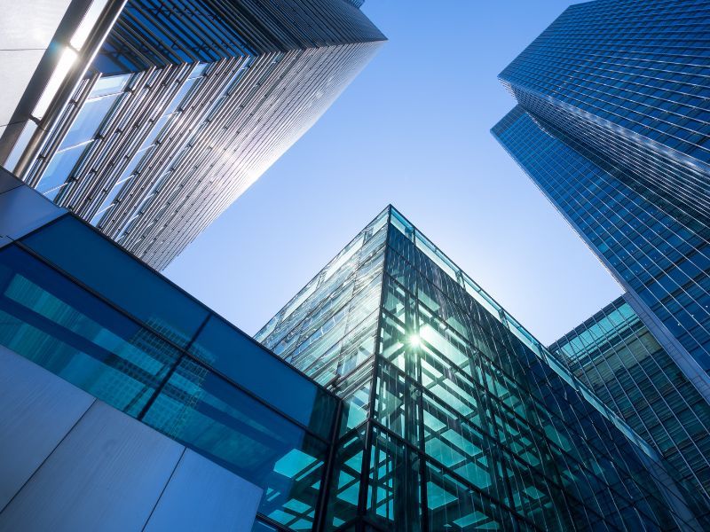 Black man in glasses wearing business clothes looks at a phone in front of a building with a bar graphing showing upward growth behind him.